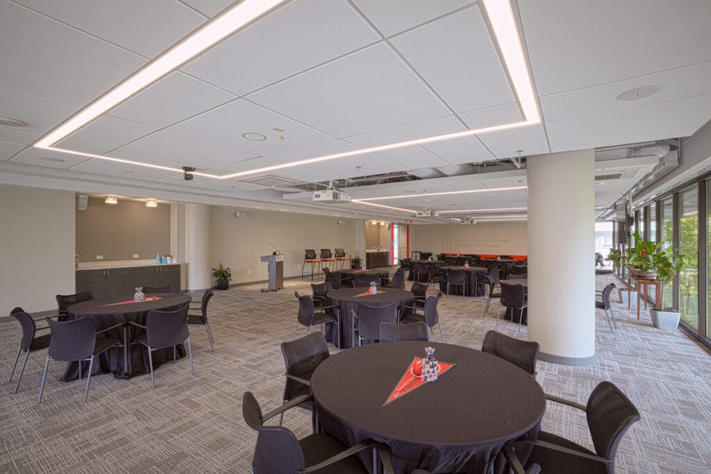 TEH_03 An expansive conference room naturally lit by glass curtain walls; large squares outlined by fluorescent lights repeat from the font to back of the room in a one column grid; round tables with 5 chairs each are placed neatly throughout the space.