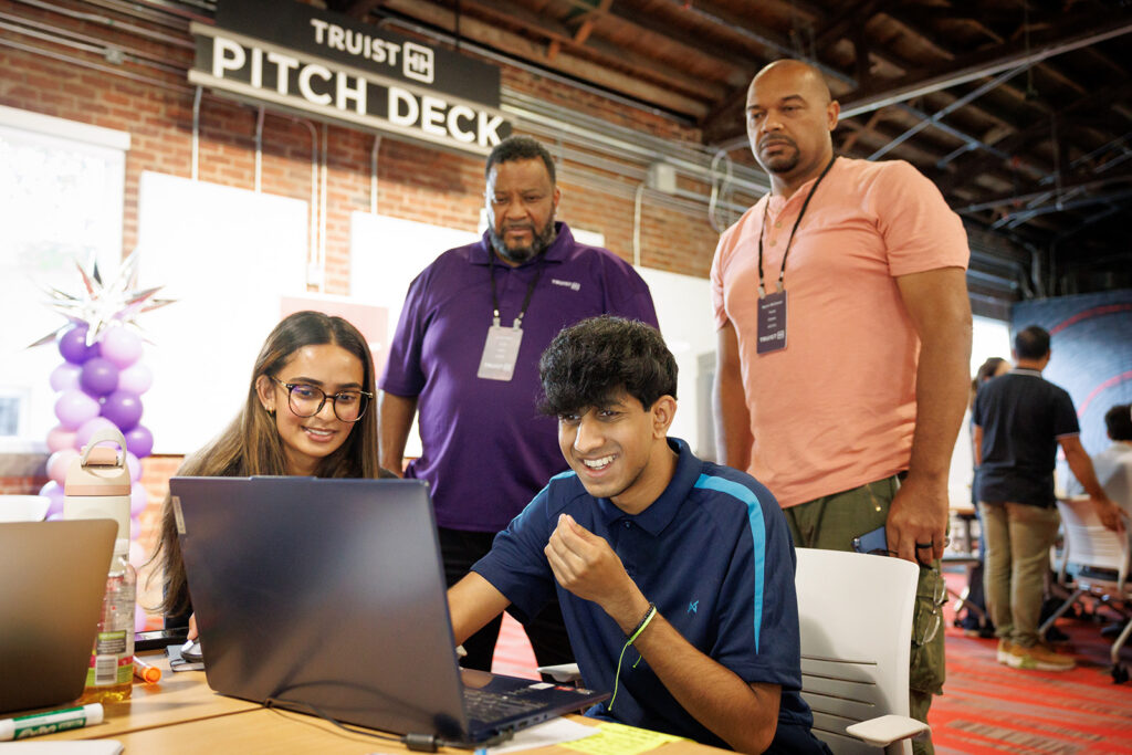 IT professionals from Truist coach two students through a problem during Students work on their computers, huddled around at table during a immersive learning experience created and hosted by Truist.. IT professionals from Truist coach two students through a problem during Students work on their computers, huddled around at table during a immersive learning experience created and hosted by Truist..