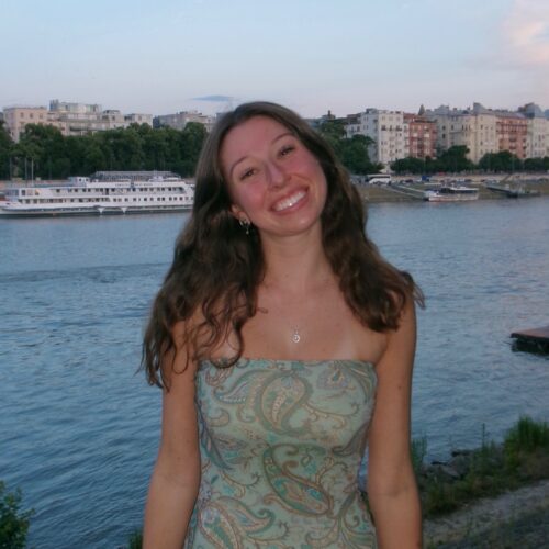 Chloe Waldron at Marg Island. She is a burnette young woman wearing a green paisley dress standing in front of a riverfront with buildings behind it.