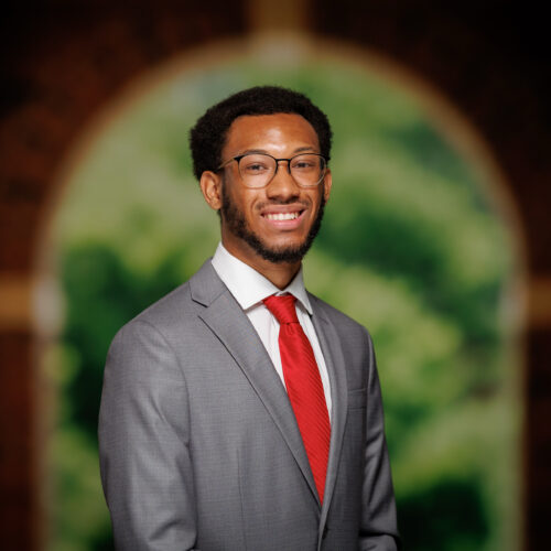 A person in a gray suit and red tie smiles while standing in front of a blurred arched background.