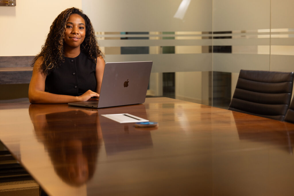 A woman smiles for a photo while sitting a table in front of a MacBook laptop.