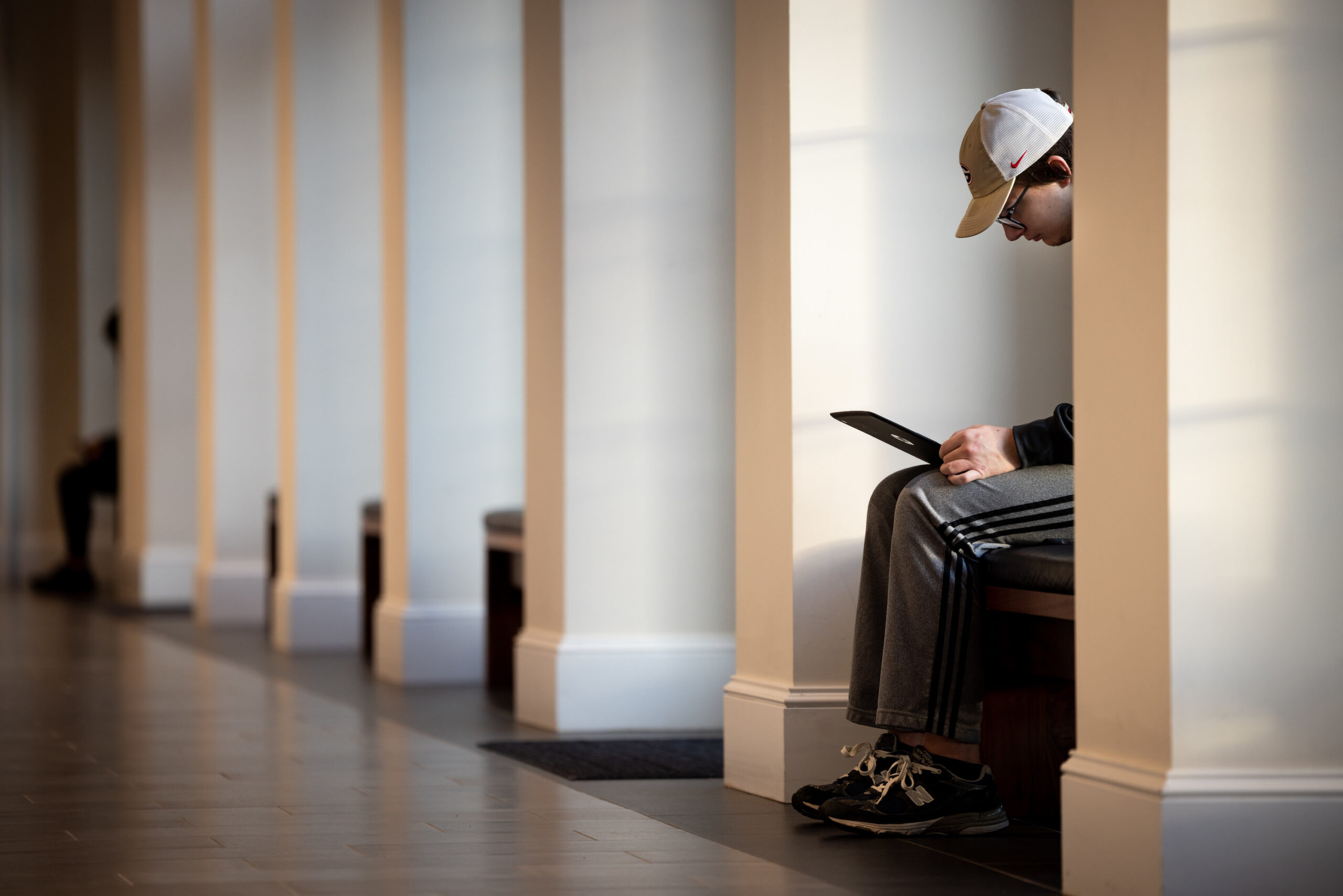 A male student sits on bench in a hallway while looking at his laptop.