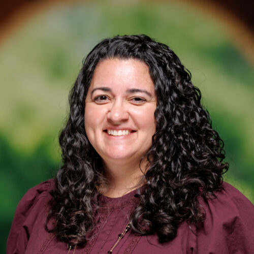 A person with long curly dark hair smiles warmly while wearing a maroon blouse and layered necklaces, standing in front of a blurred arched background.