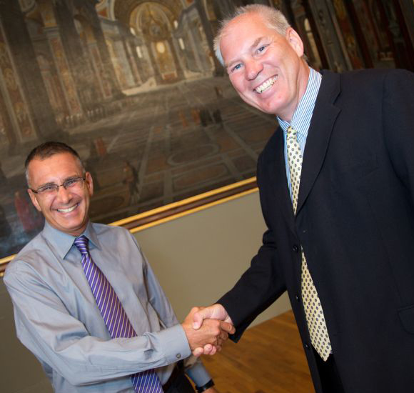 Two men in professional attire shaking hands and smiling at camera