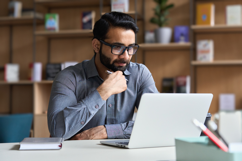 A man with glasses and a beard holds a pen as he looks at a laptop at his desk.