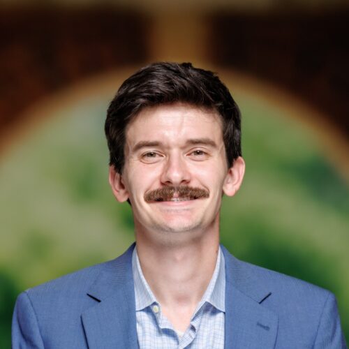A person with a mustache wears a blue blazer over a checkered shirt, standing in front of a blurred arched doorway with greenery visible behind.