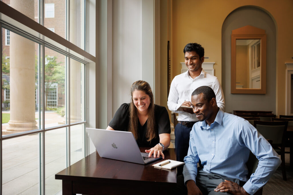 Two men and a woman smile while looking at a laptop on a desk.