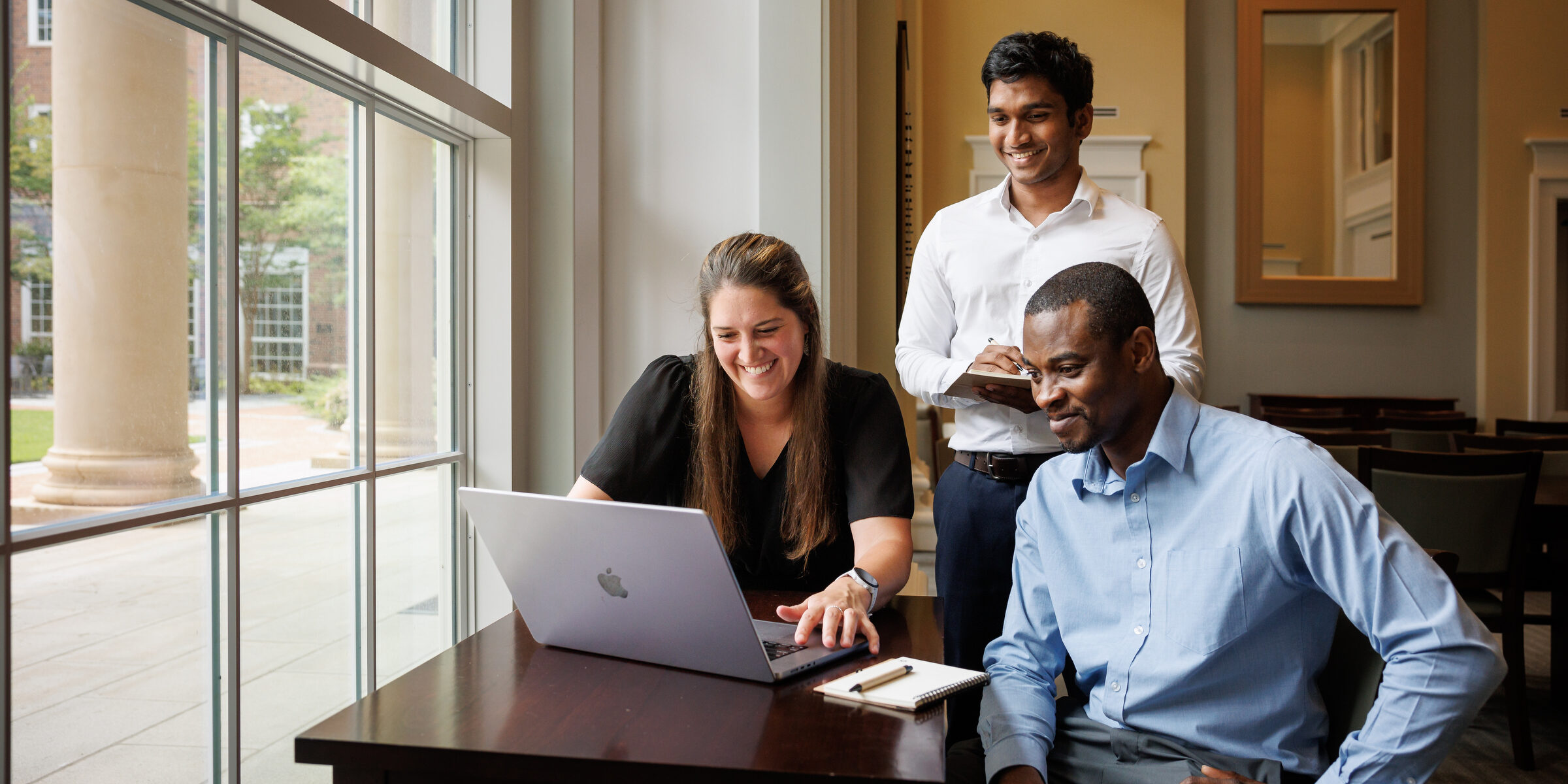 Two men and a woman smile while looking at a laptop on a desk.