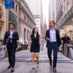 three young adults walk in front of the New York Stock Exchange in business attire