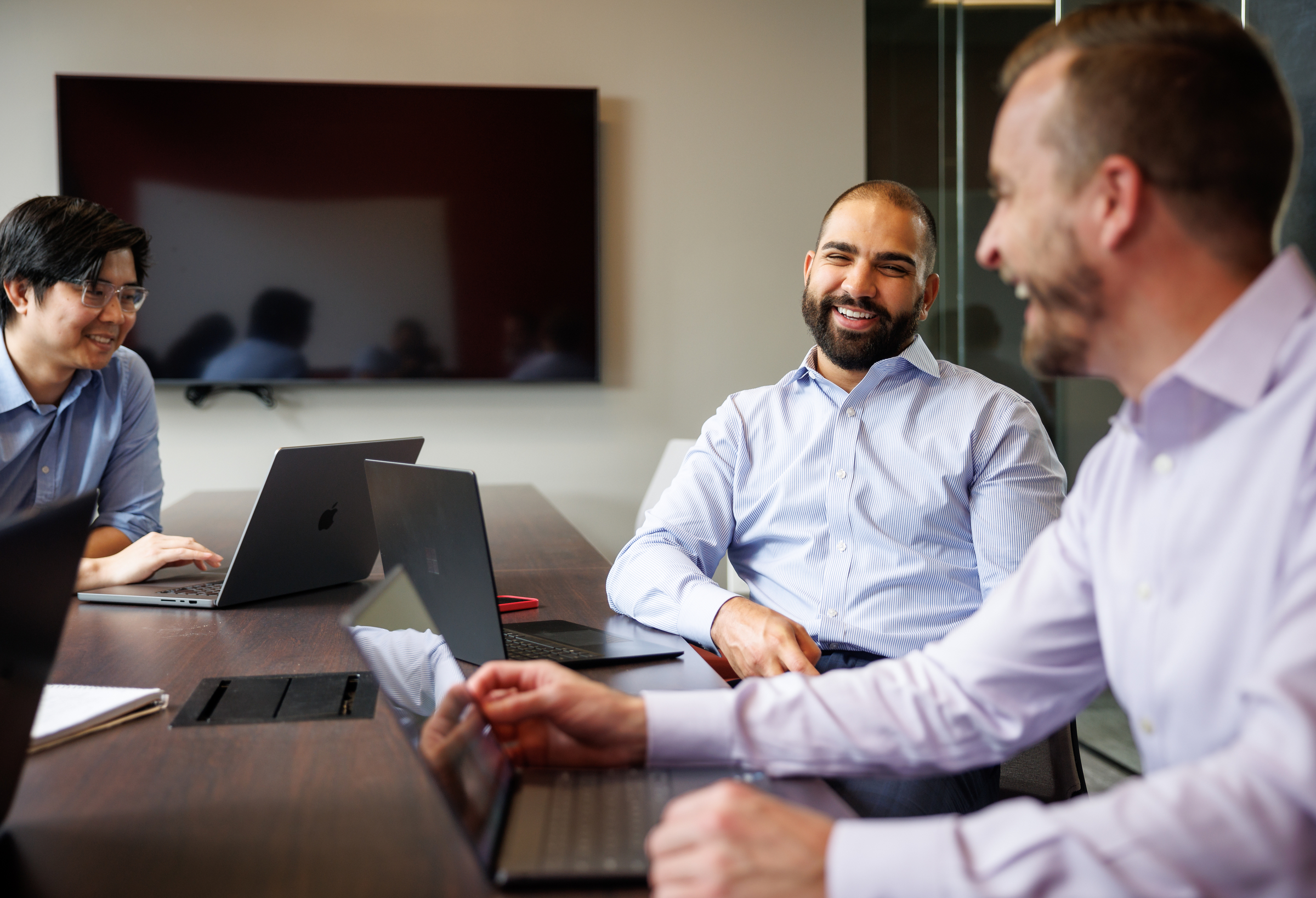 Three men with laptops laugh while talking at a table in a conference room.