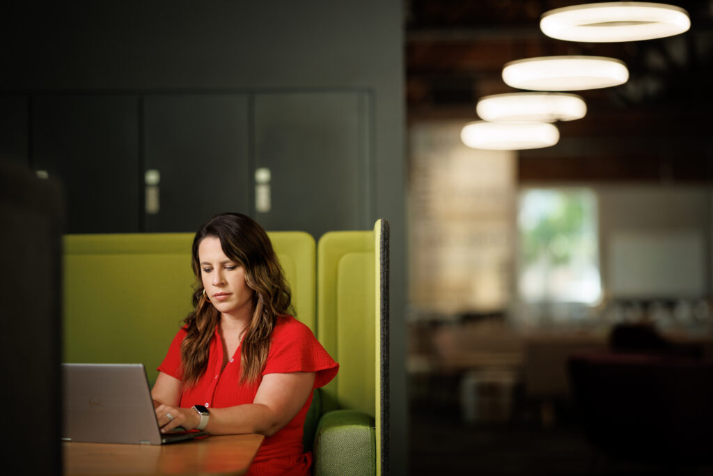 A woman sits at a booth while looking at a laptop in a dimly-lit room,