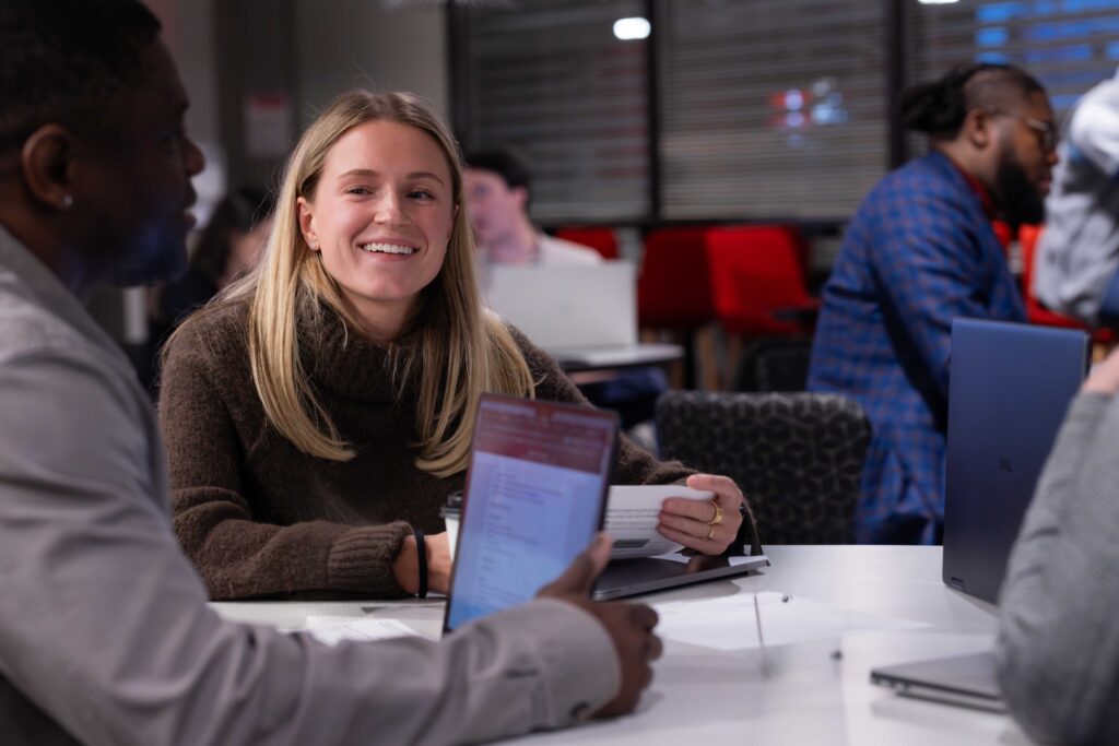 pmba-class A female Professional MBA student smiles at a table while talking to a male Professional MBA student with a laptop.