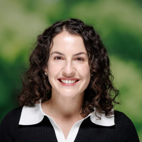 A person with curly dark hair smiles warmly while wearing a black ribbed top with a white collar and cuffs, standing in front of a blurred green and brown background.