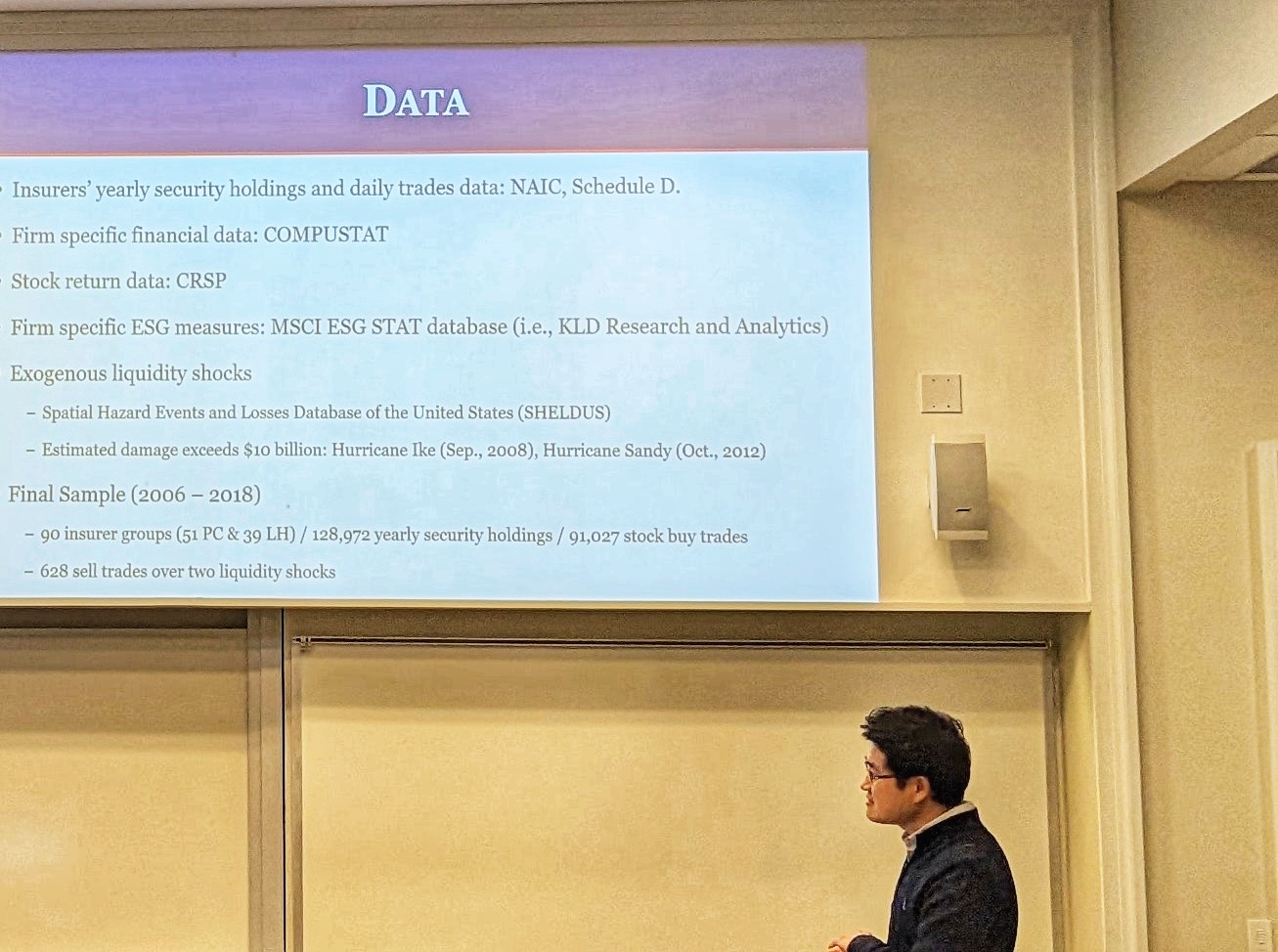 A male speaker talks to the audience in front of a presentation screen during a Risk Management & Insurance Symposium in a BLC classroom.