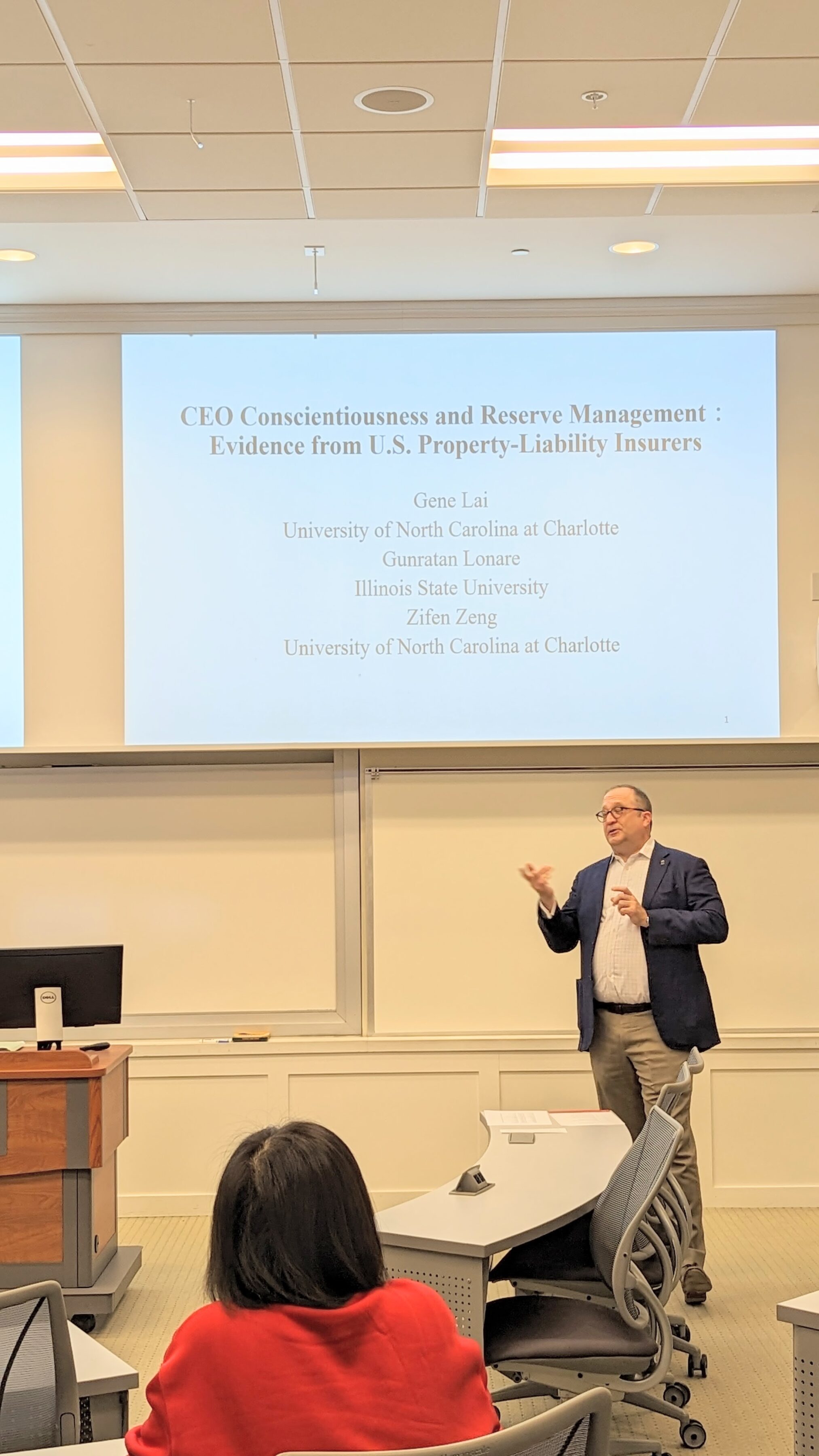 A male speaker talks to the audience in front of a presentation screen during a Risk Management & Insurance Symposium in a BLC classroom.