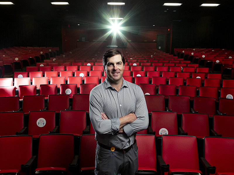 John Rooker stands arms crossed in front of a row of red movie theater seats
