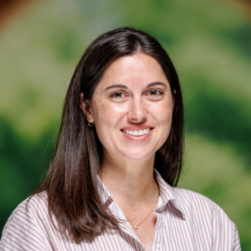 A smiling person with long dark hair stands in front of a blurred green and brown background, wearing a striped short-sleeved shirt.