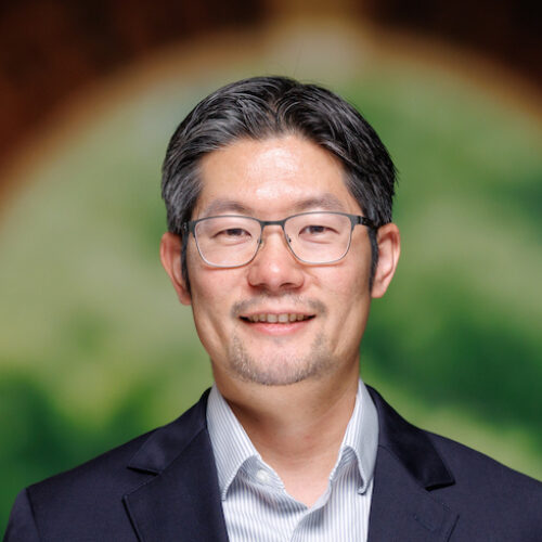 A person with glasses and a dark suit stands in front of a blurred arched background featuring green foliage.