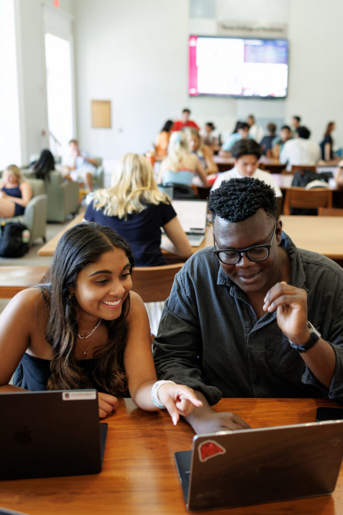 Three students collaborate at a wooden table in a bright, bustling library or study hall, surrounded by other learners and large windows.