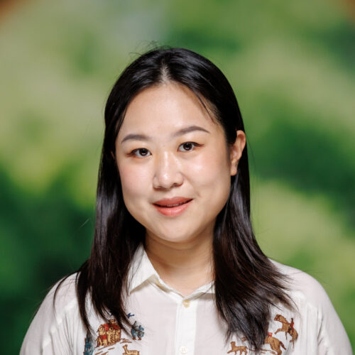 A person with long dark hair stands in front of a blurred green and brown archway, wearing a light-colored shirt with embroidered designs.
